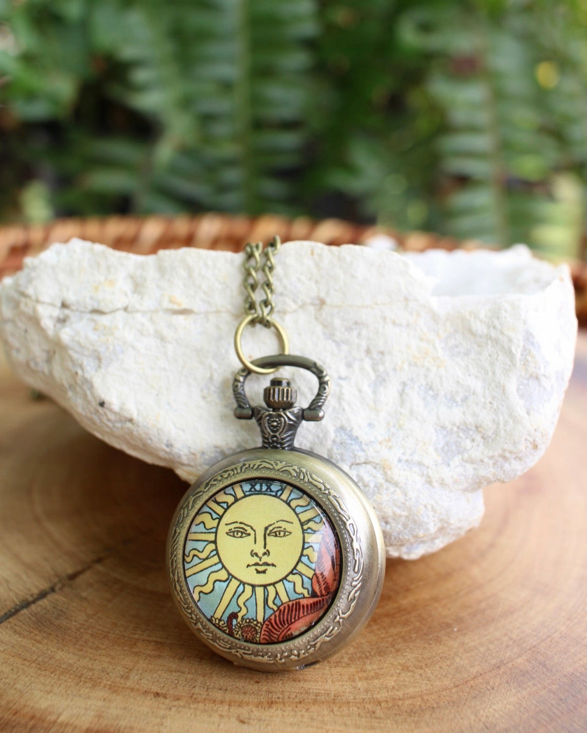 A pocket watch necklace with an antique bronze finish and a sun face illustration, resting on a white stone over a wooden surface with green foliage in the background.