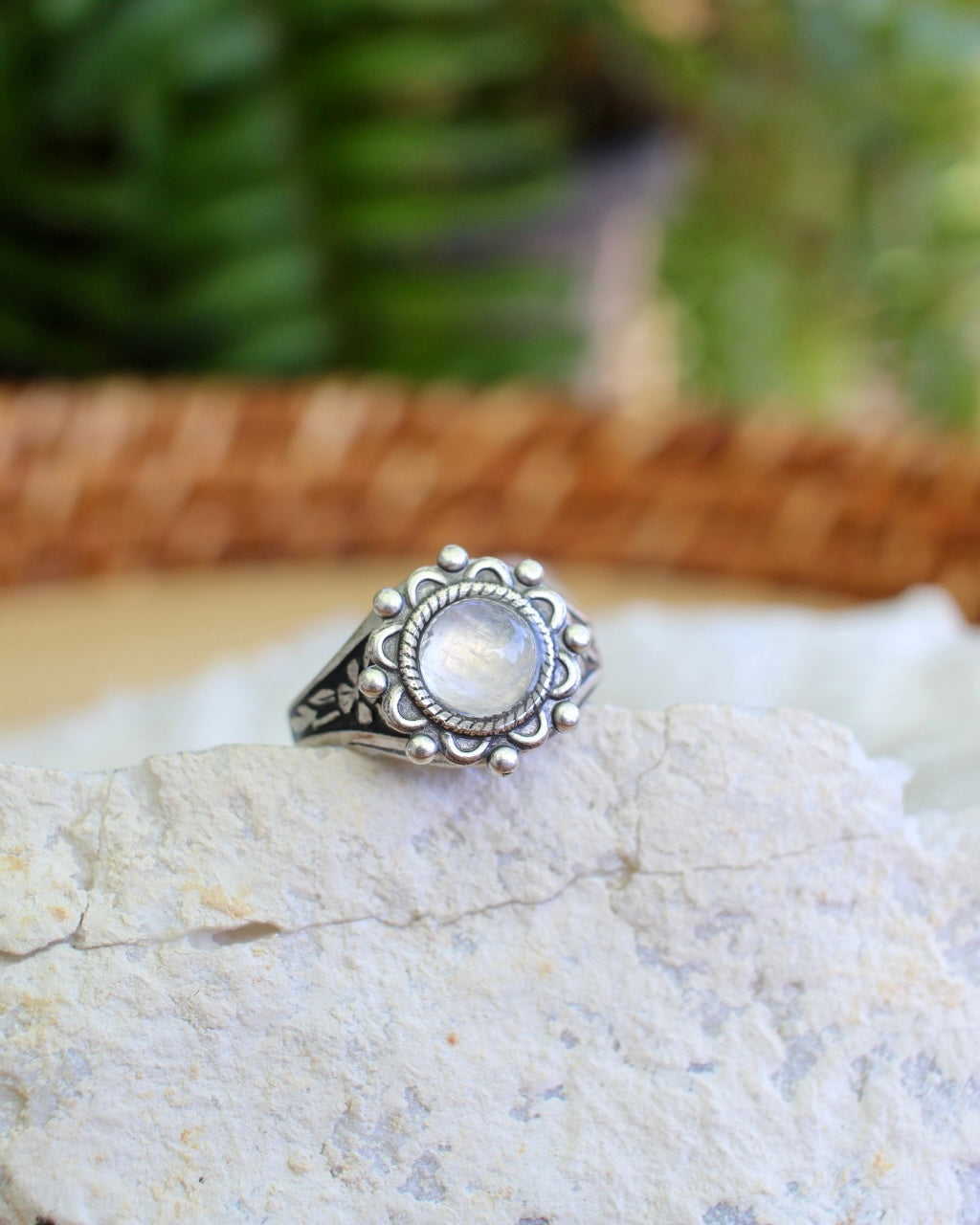 A silver ring with a round, milky white moonstone gemstone in an ornate floral setting, displayed on a textured white stone with a blurred green and brown background.