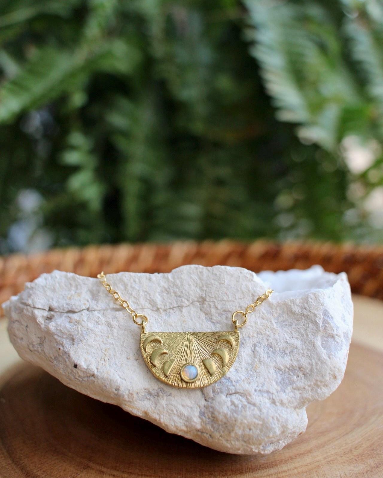Gold necklace with a half-circle pendant with moon phases on a white stone with green foliage in the background