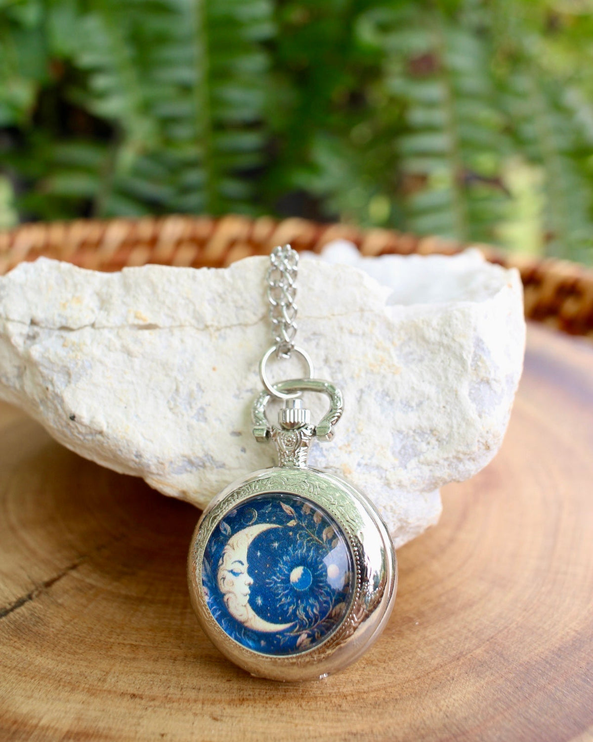 A silver-toned pocket watch pendant necklace with a blue and white celestial design, resting on a white stone over a wooden surface with a blurred green fern background.