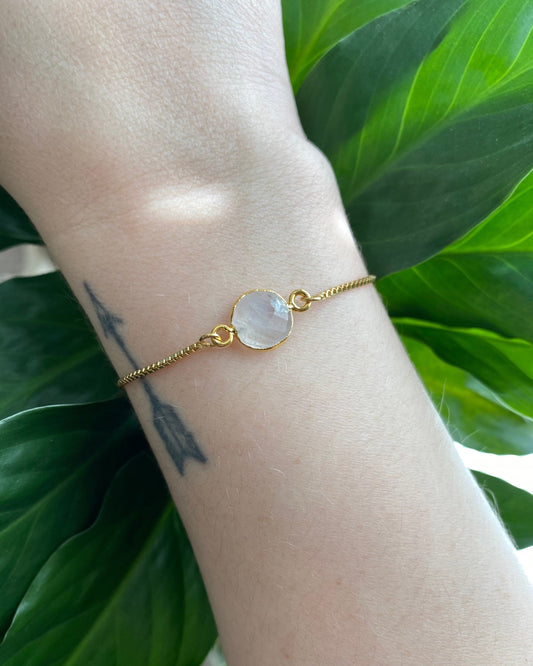 Gold bracelet with a stone on a wrist against a green leafy background