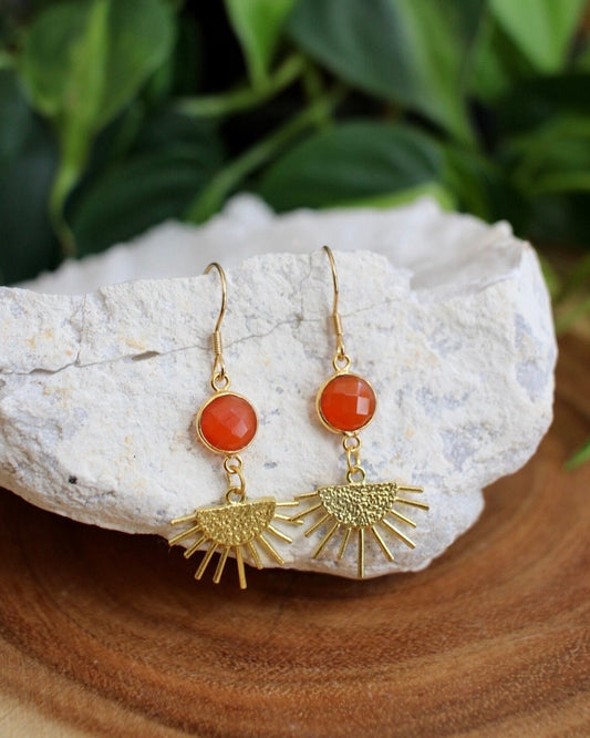 Gold earrings with red orange stones on a white stone surface with green leaves in the background