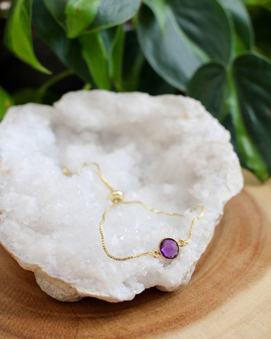 Gold bracelet with a purple gemstone on a white stone with green leaves in the background