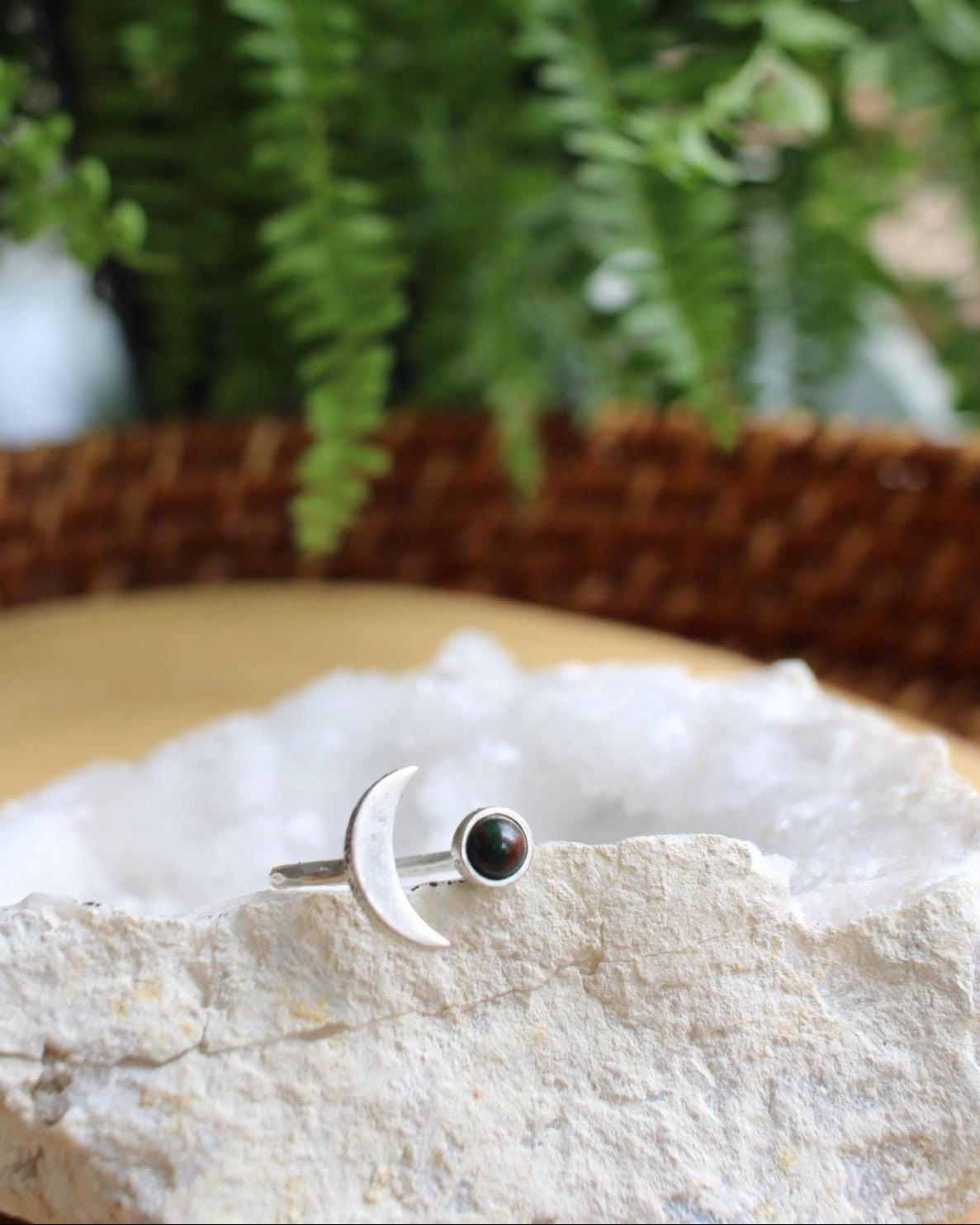 Silver ring with a crescent moon on one side and a bloodstone on the other sitting on a white crystal with green fern in the background