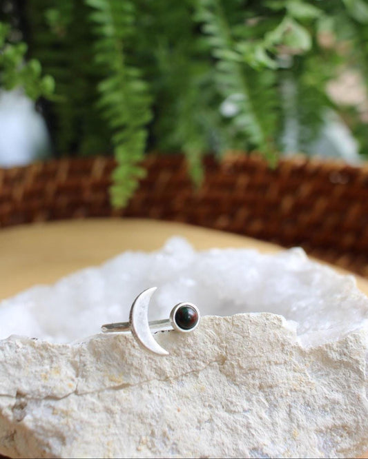 Silver ring with a crescent moon on one side and a bloodstone on the other sitting on a white crystal with green fern in the background