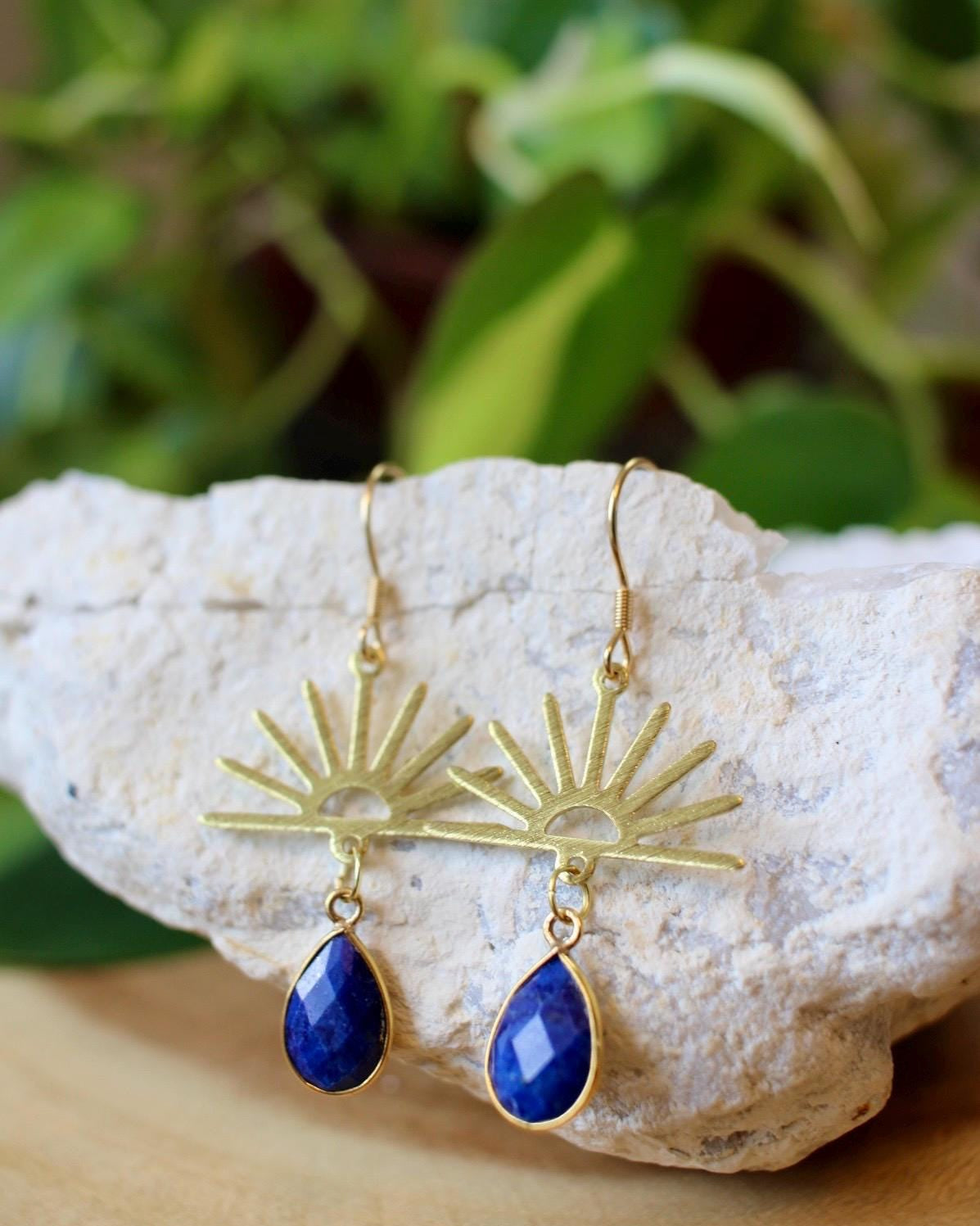 Gold earrings with blue teardrop stones on a rock with green foliage in the background