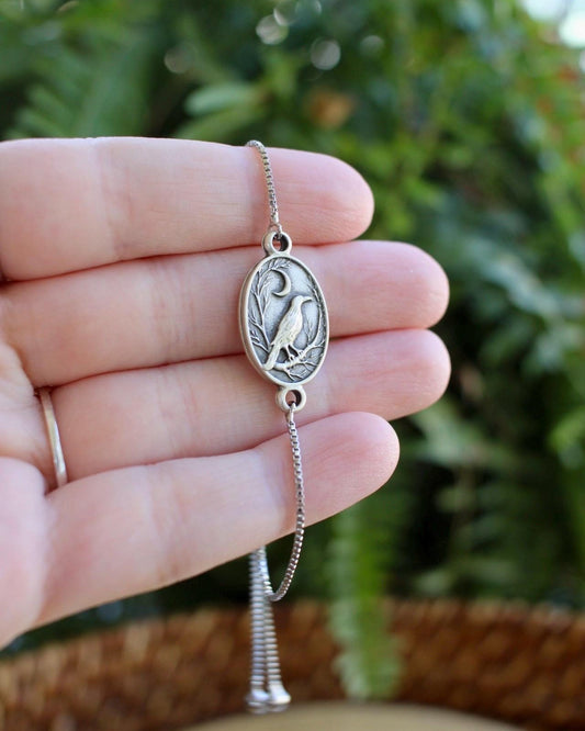 Silver bracelet with a raven and moon charm held in a hand against a blurred green background