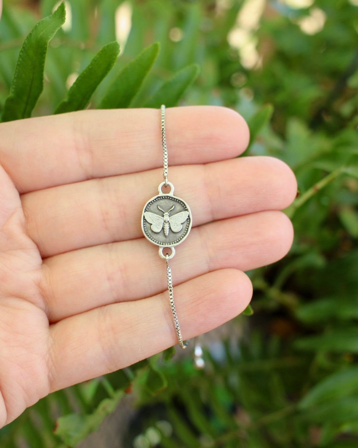 A delicate silver-toned chain bracelet with a moth charm, held in a person's hand against a blurred green garden background.