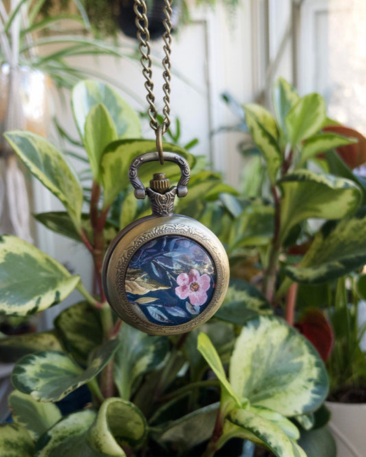 Decorative pocket watch necklace with floral design hanging among plants