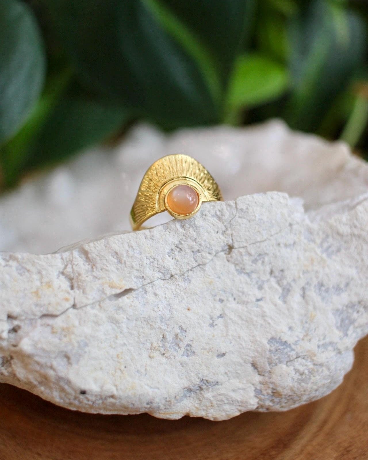 Gold ring with a peach stone on a rock with green foliage in the background