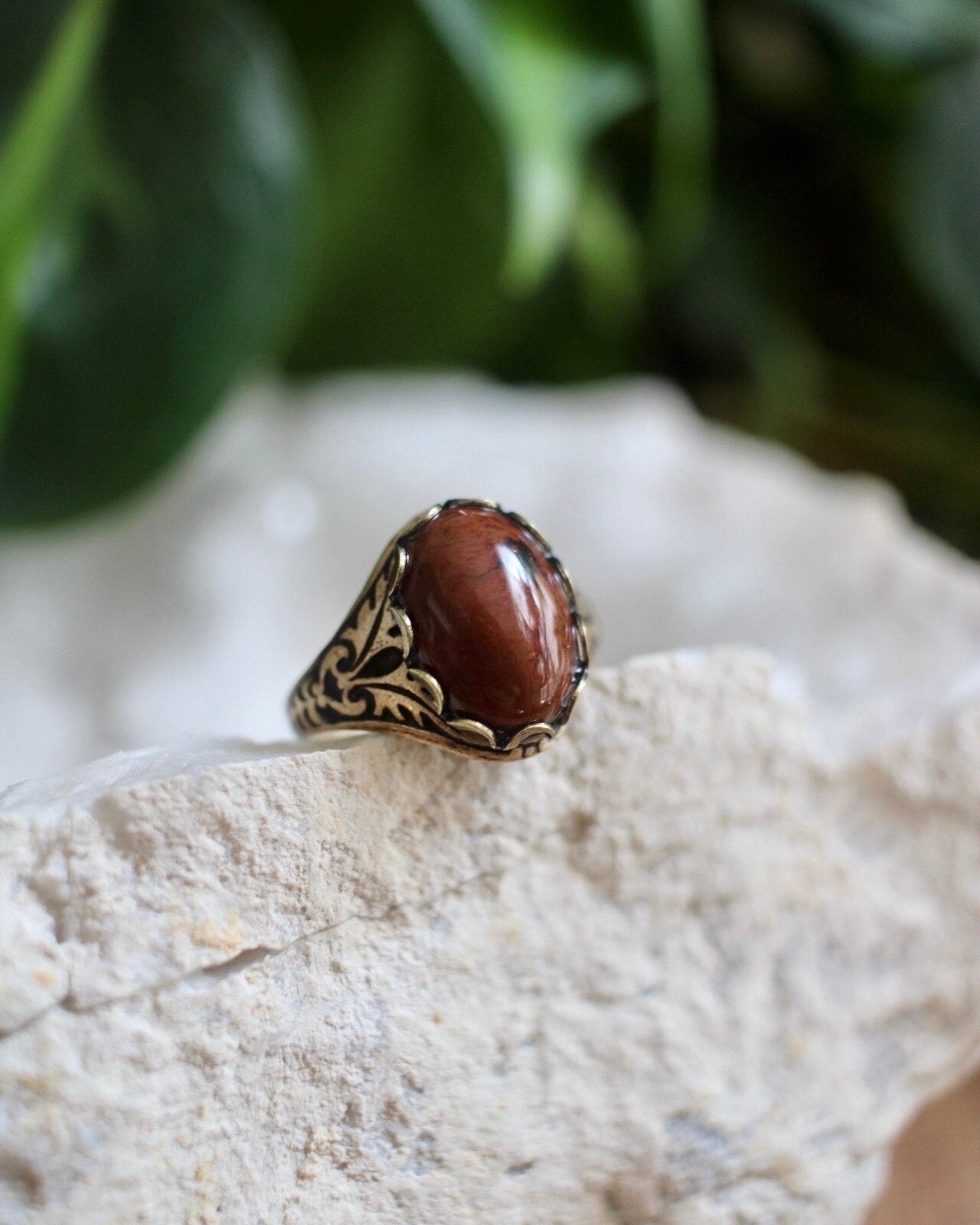 Decorative ring with a reddish brown stone on a white rock with green foliage in the background