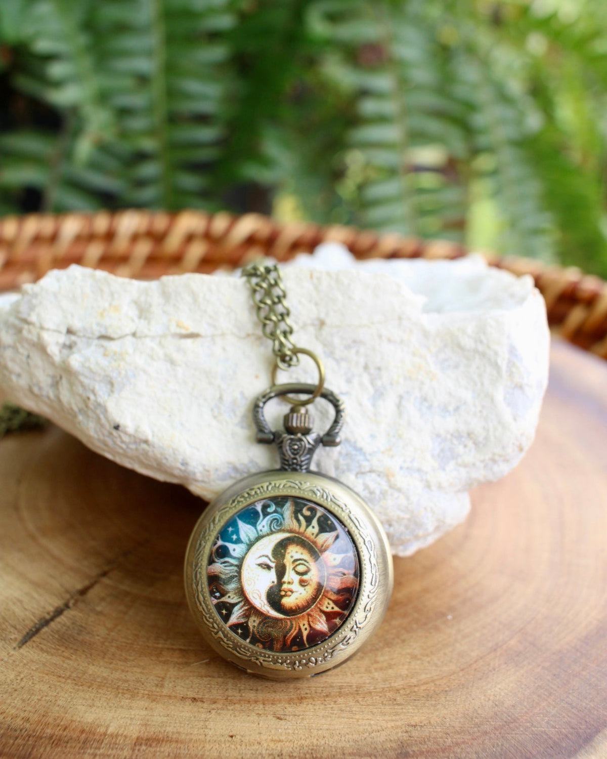 A round vintage-style pendant necklace with an antique bronze frame and a colorful sun-and-moon illustration, resting on a white stone over a wooden surface with green foliage in the background.
