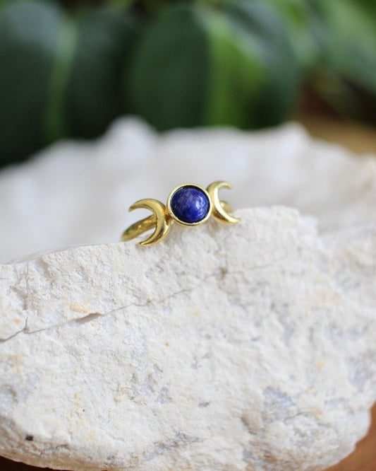 Gold ring with a blue stone on a white stone surface with green leaves in the background