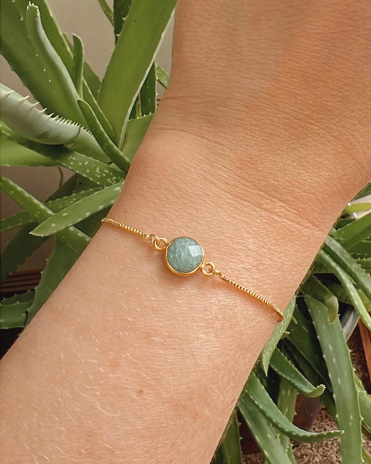 Gold bracelet with a green gemstone on a wrist against a plant background