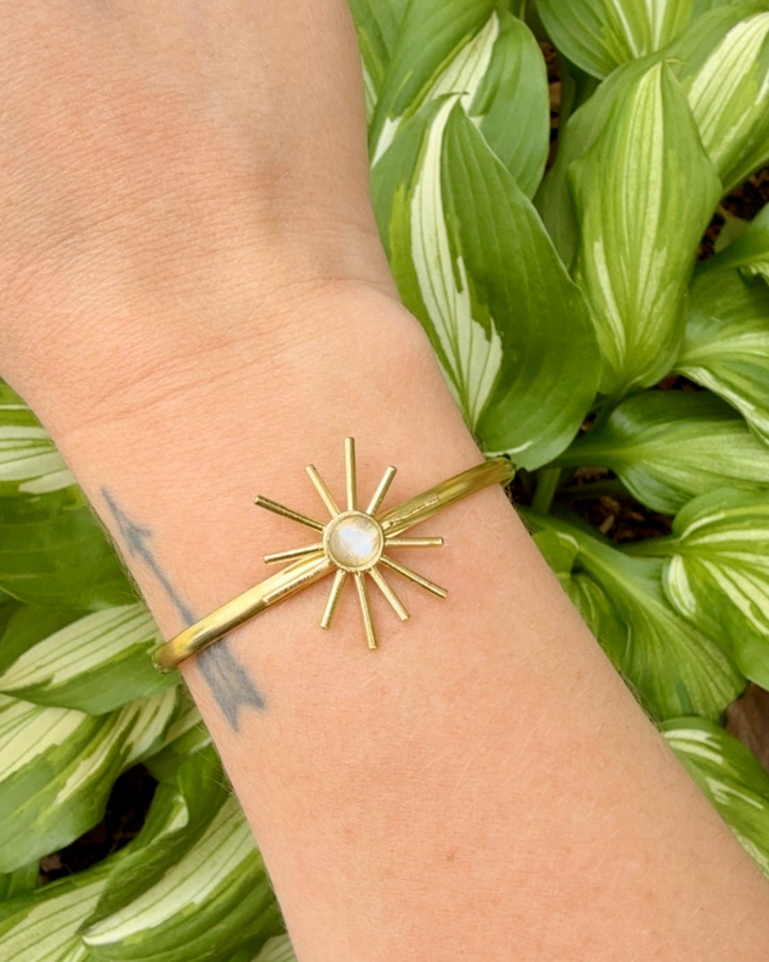 Gold bracelet with a sun design and moonstone in center worn on a wrist against a green leafy background