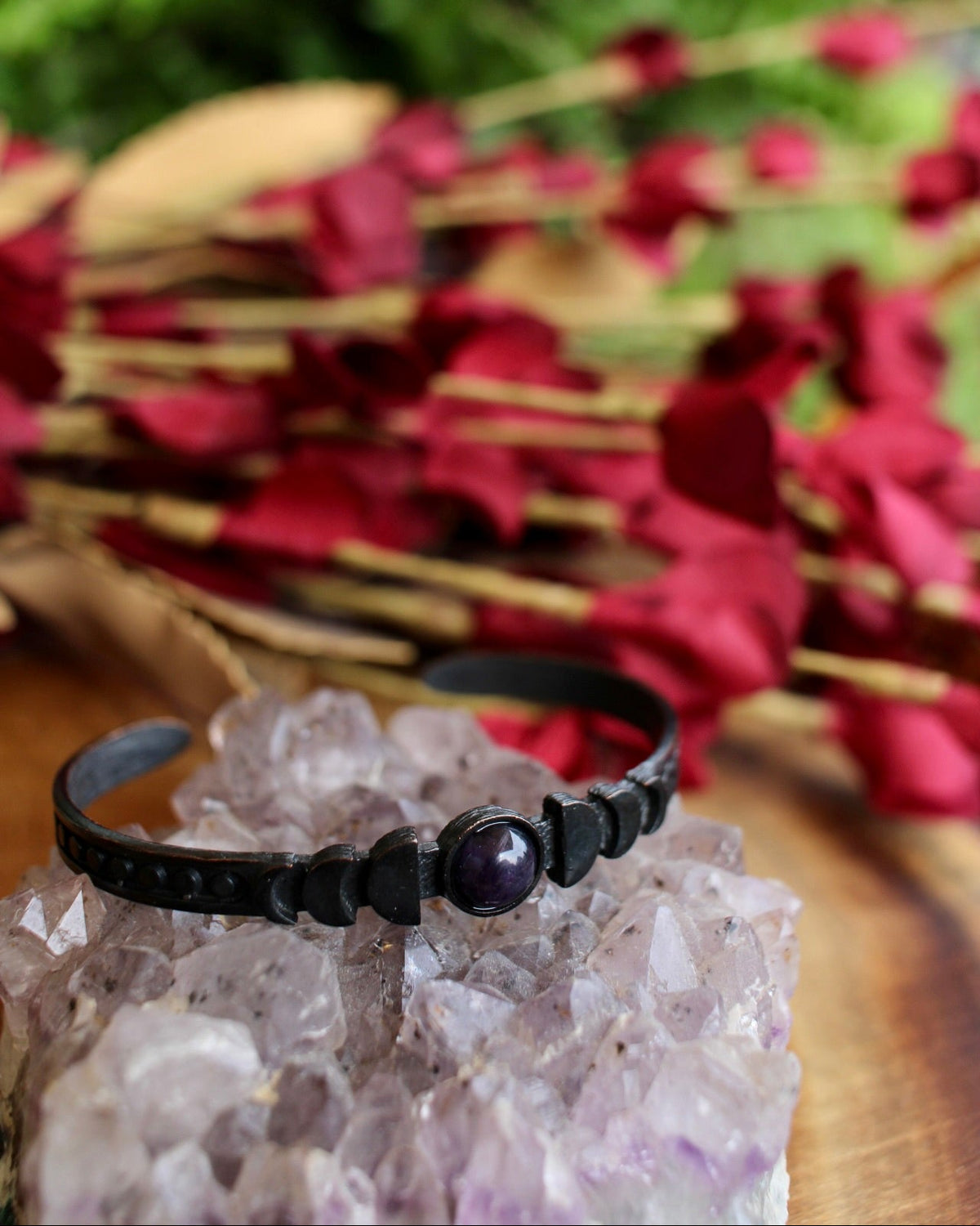 A black moon phase cuff with an amethyst stone in the center sitting on a raw amethyst stone with a flower bouquet in the backgroud