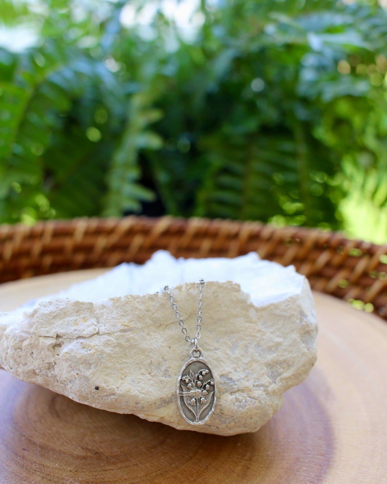 A silver oval pendant necklace with a floral design hangs from a chain and is displayed on a white stone, set on a wooden surface with a blurred green plant background.