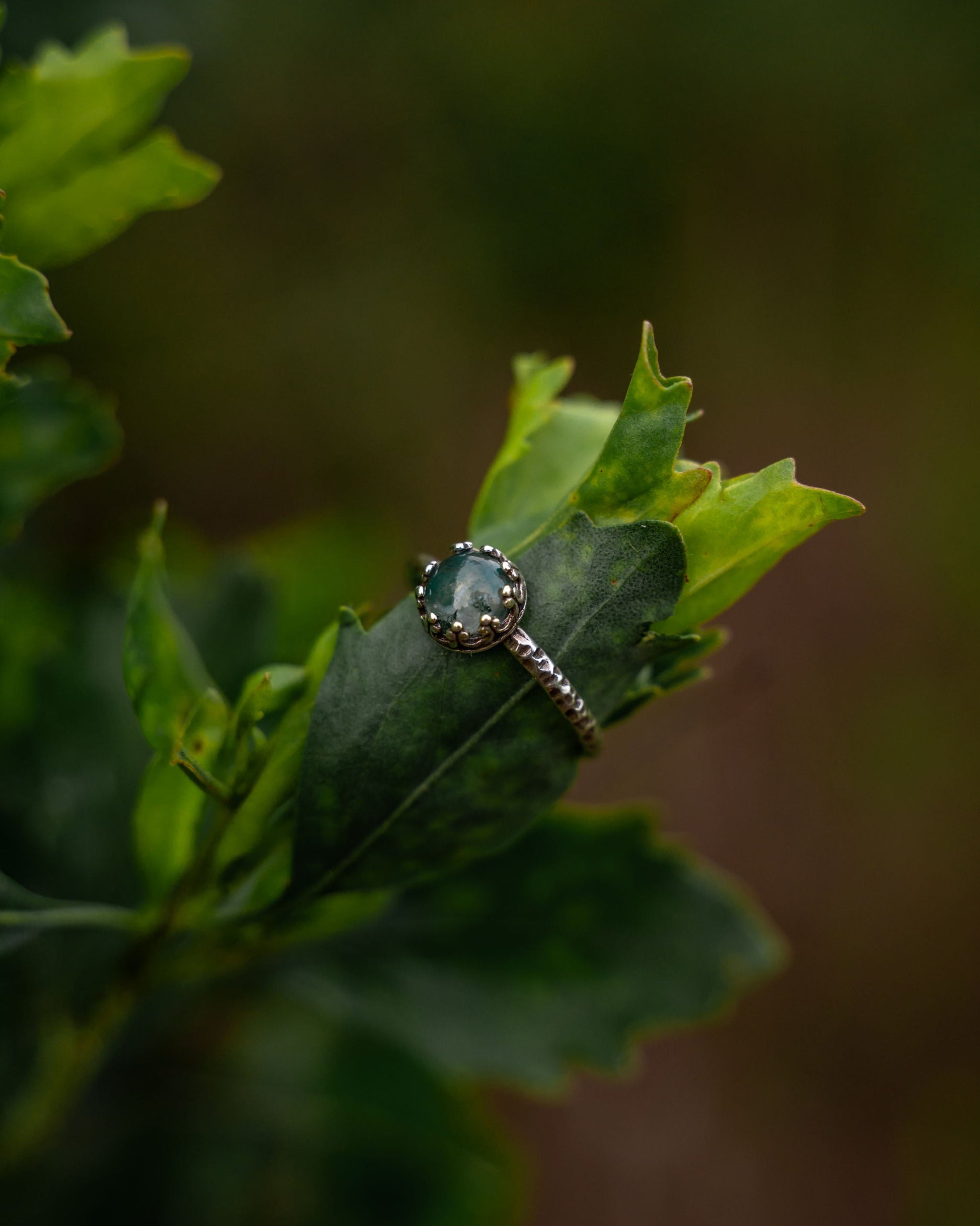 A silver-toned ring with a round green gemstone and small clear accents, resting on a green leaf against a blurred natural background.