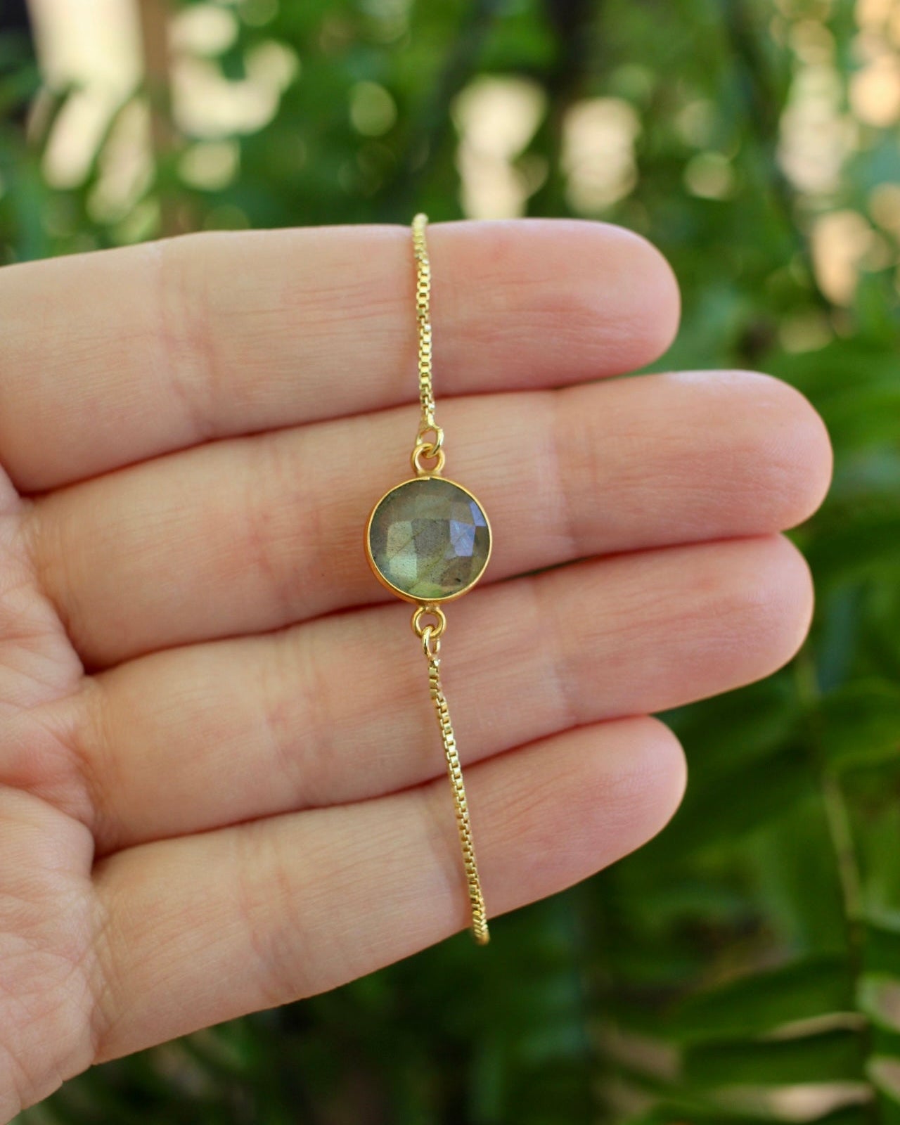 Green stone bracelet one a gold chain held in a hand infront of green plant