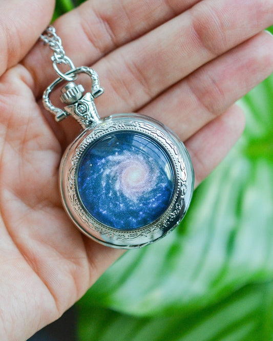 Silver pendant with a blue galaxy design being held in a hand in front of a green plant