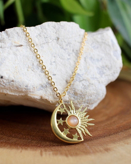 Gold necklace with a moon and sun pendant on a rock with greenery in the background