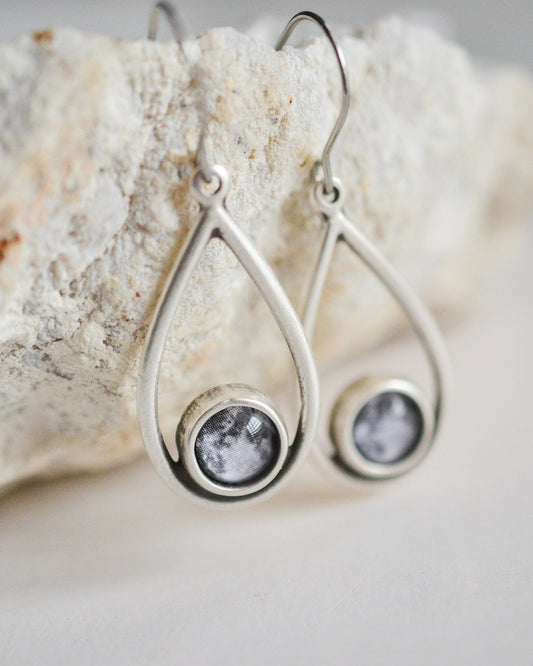 Silver teardrop earrings with full moon prints hanging from a textured stone surface