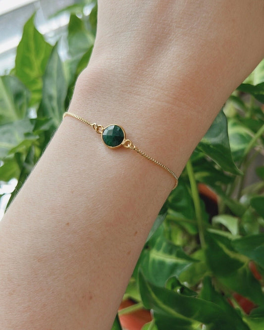 Gold bracelet with a green gemstone on a wrist against a blurred green leafy background