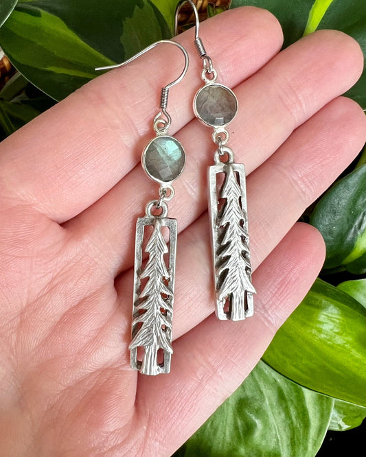 Pine tree dangle earrings with blue green stones laying on a hand in front of a plant