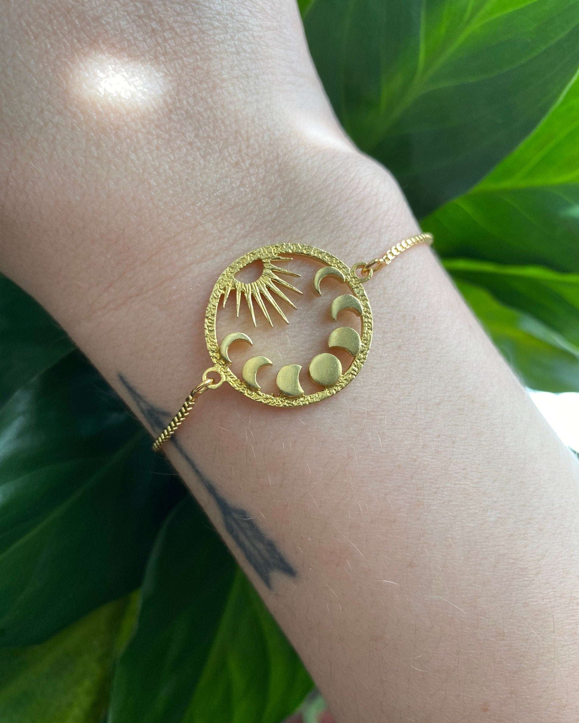 Gold bracelet with sun and moon design on a wrist against a green leafy background