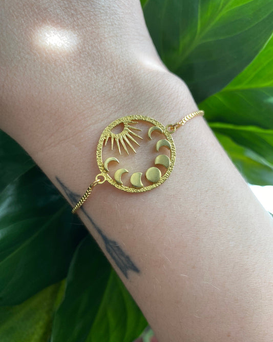 Gold bracelet with sun and moon design on a wrist against a green leafy background