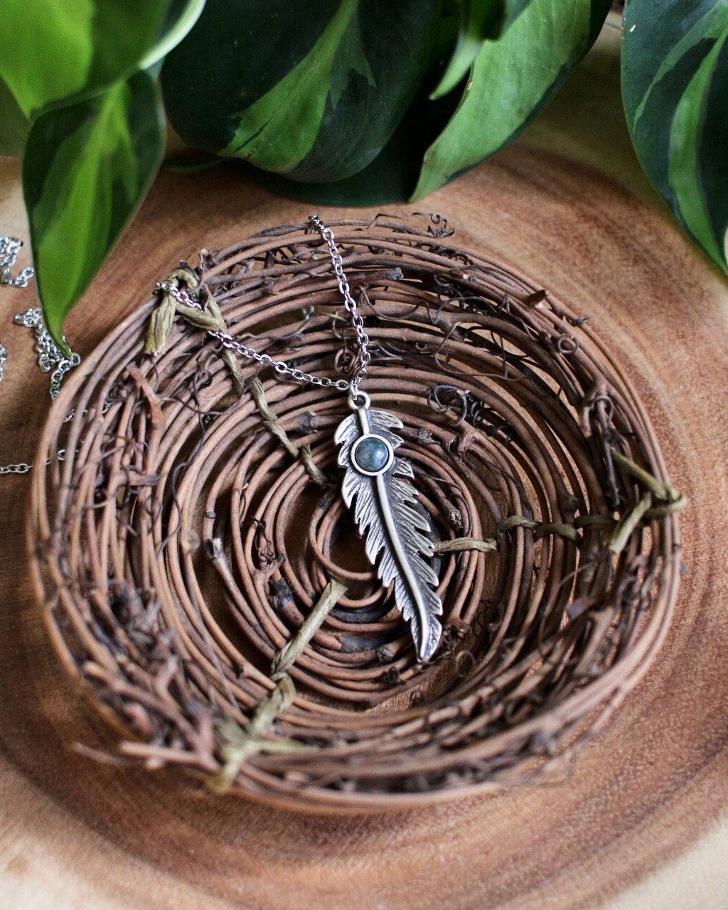 Silver feather necklace on a wooden surface with green leaves in the background